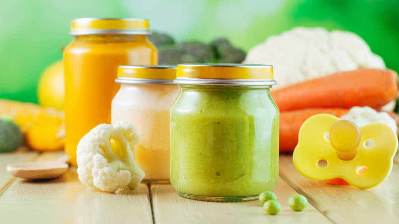 papinhas para bebês em potes de vidro, uma papinha verde e duas amarelas. Ao lado, uma chupeta amarela, ao redor, vários legumes.