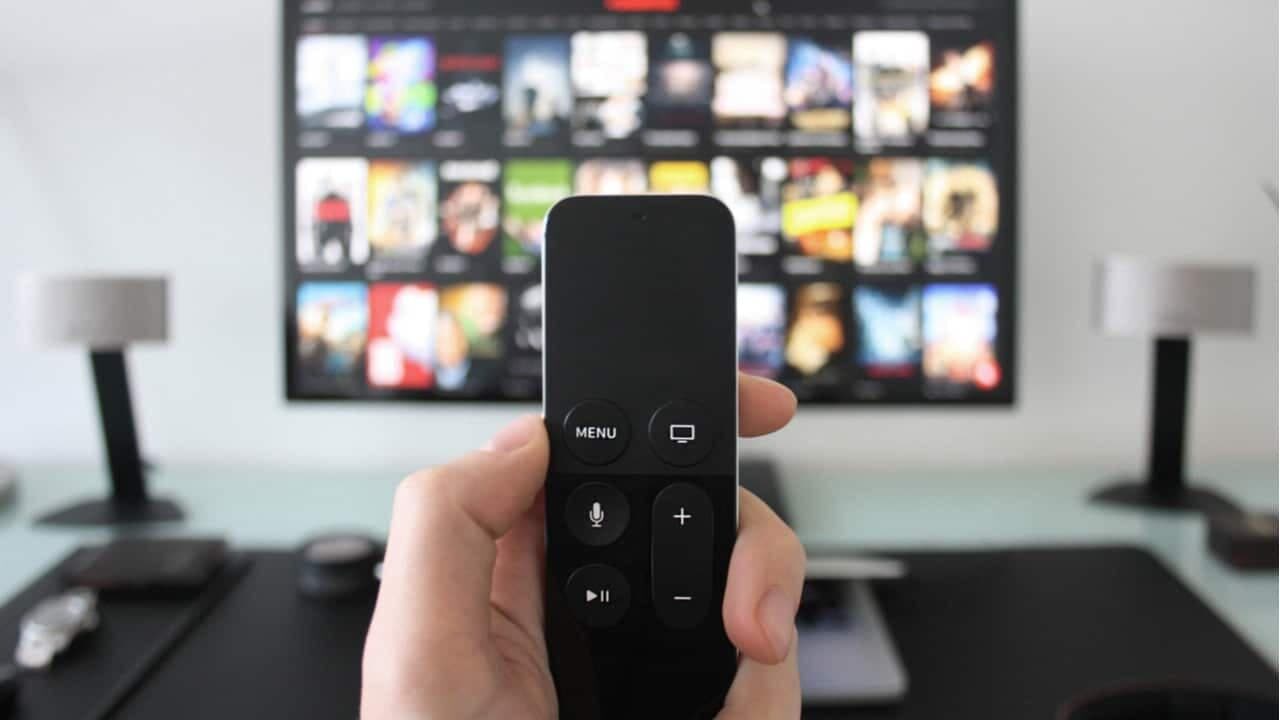 Mão segurando controle remoto e TV ao fundo mostrando catálogo de filmes ou séries