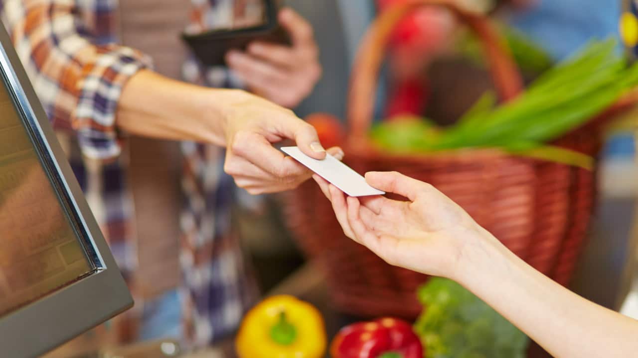Mão de uma pessoa entregando cartão de vale-alimentação na mão de outra. Ao fundo, alguns legumes e verduras aparecem desfocados.