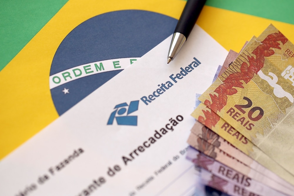 Uma parte da bandeira do Brasil, somente até a frase "ordem", coberta por uma folha escrita "Receita Federal" e notas de 20 reais, além de uma caneta. reforma tributária