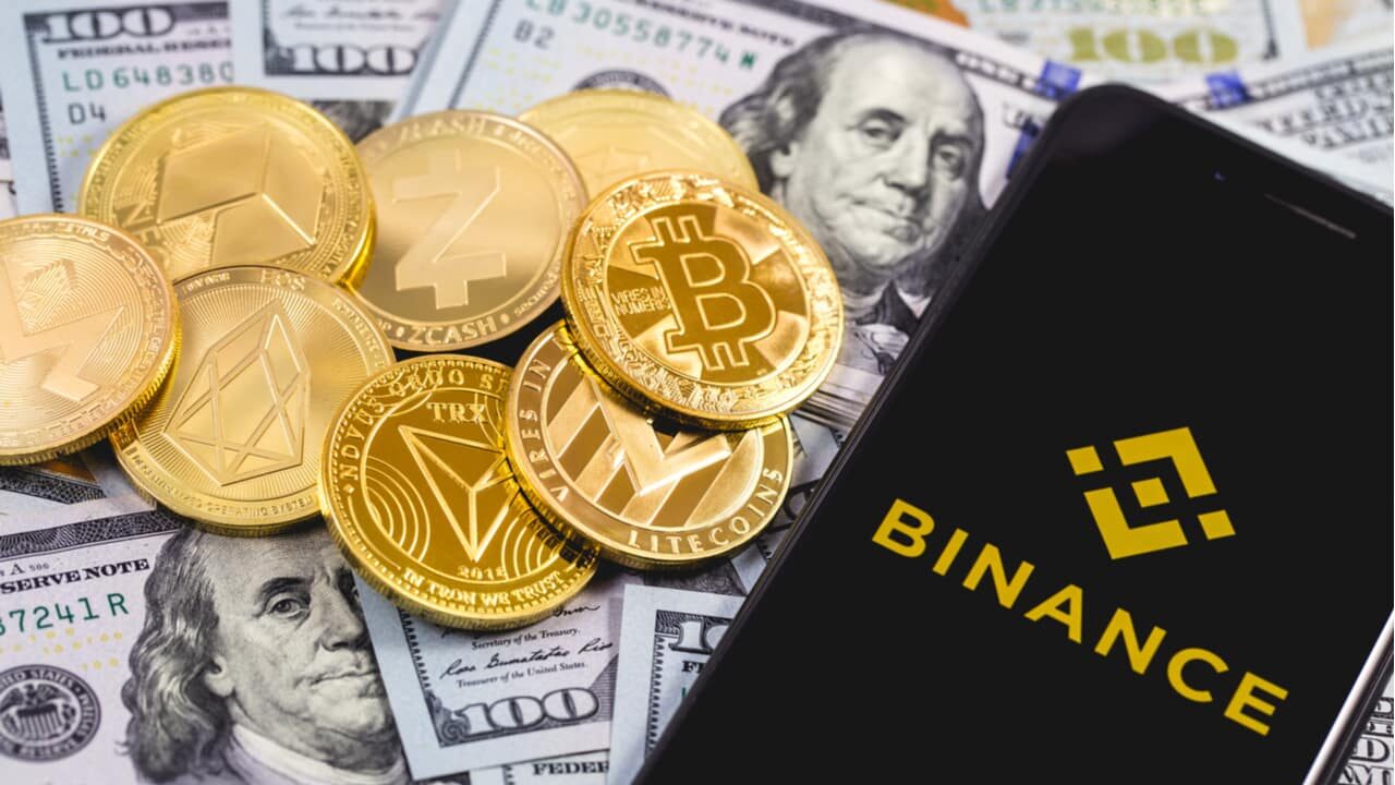 Celular com a logo da Binance na tela, ao lado de várias criptomoedas e por cima de notas de dólares