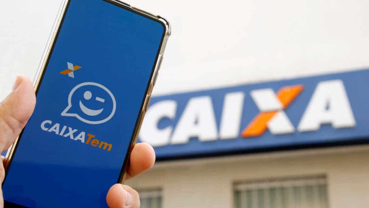 Celular na mão com app Caixa Tem aberto na tela inicial. Ao fundo, uma agência da Caixa Econômica Federal.