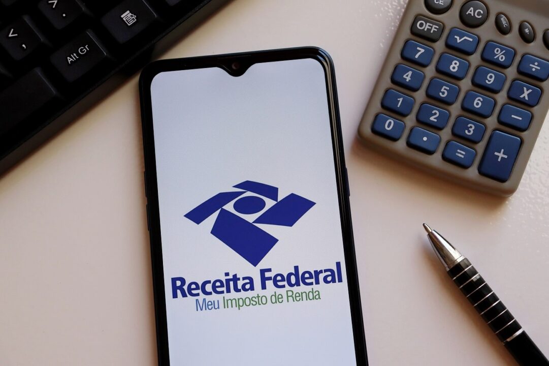Na imagem aparece um celular com a logo da Receita Federal na tela, uma calculadora, uma caneta e parte do teclado de um computador.
