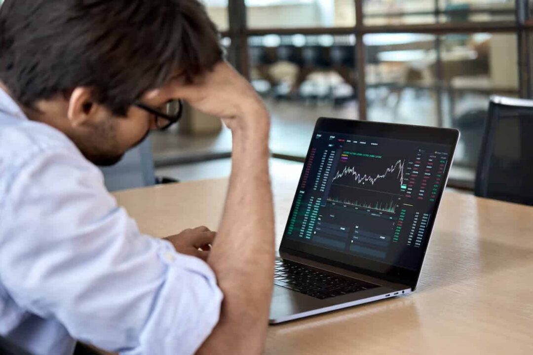Imagem de homem cabisbaixo observando tela de notebook que apresenta um gráfico com índices ruins de investimento