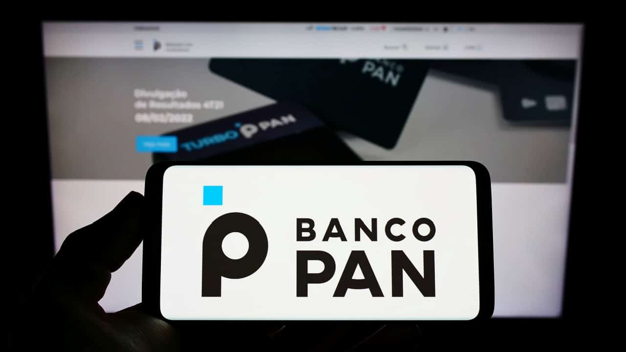 Posicionada em frente a uma tela com informações do Banco Pan, mão segura celular que exibe a logo da instituição