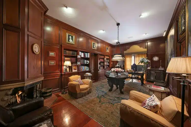 Biblioteca da antiga mansão de Madonna com poltronas, tapete, estantes e lareira. 
