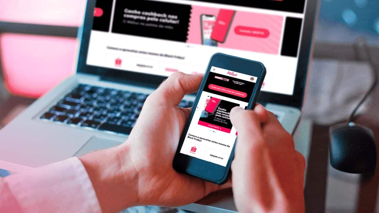 mãos usando celular e fazendo compras pelo aplicativo Méliuz com um computador aberto no site da empresa ao fundo
