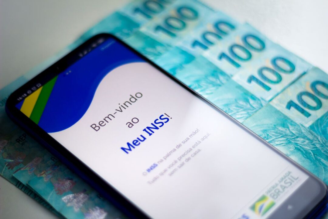 Celular sobre notas de cem reais, enquanto exibe a tela inicial do app Meu INSS