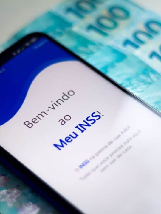Celular posto sobre notas de cem reais exibindo a tela inicial do app Meu INSS