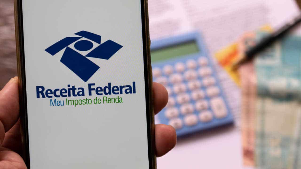 Celular na mão com app do Imposto de Renda na tela inicial, há calculadora ao fundo