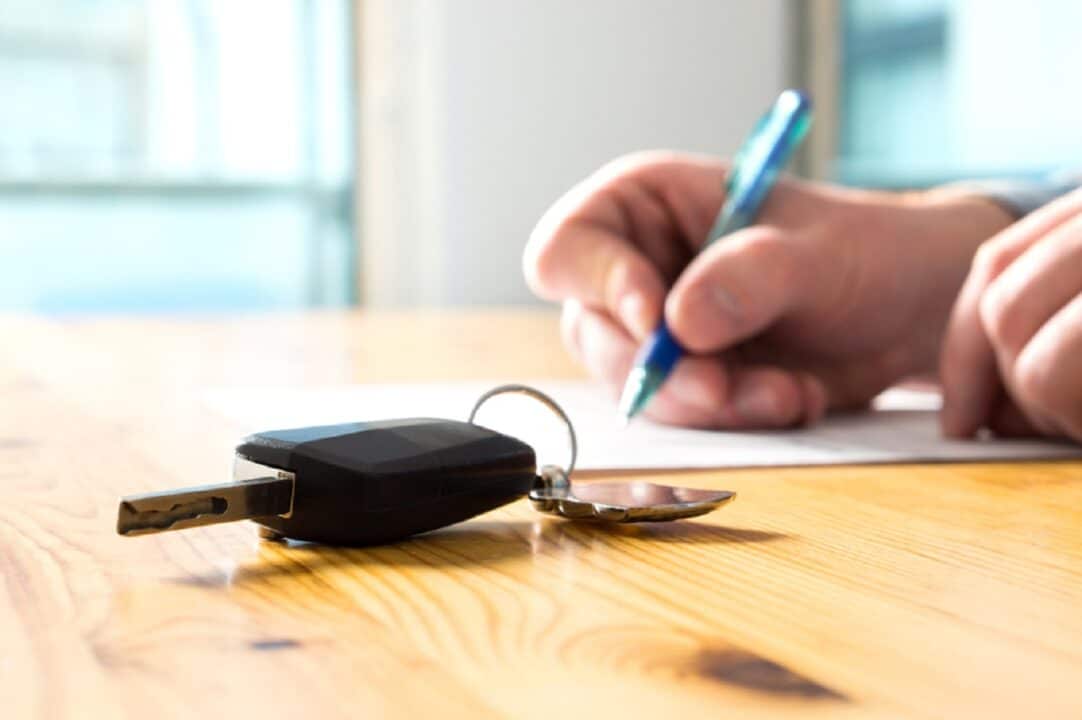 Imagem de uma mão assinando um documento que está sob uma mesa. Também sob a mesa, está a chave de um automóvel.
