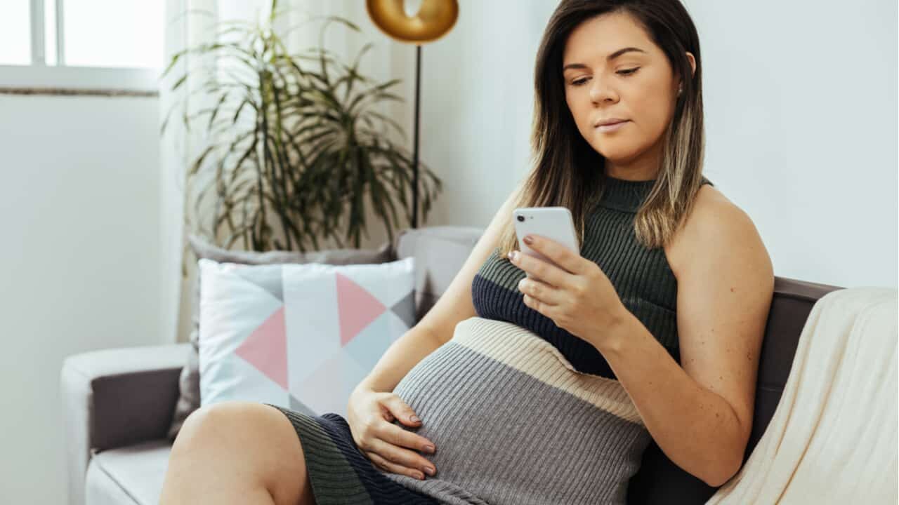 Mulher grávida sentada no sofá segurando seu celular com a mão esquerda, enquanto segura sua barriga com a mão direita.