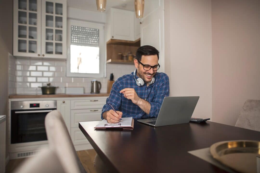Imagem mostra um homem sorrindo enquanto trabalha no computador dentro de casa.