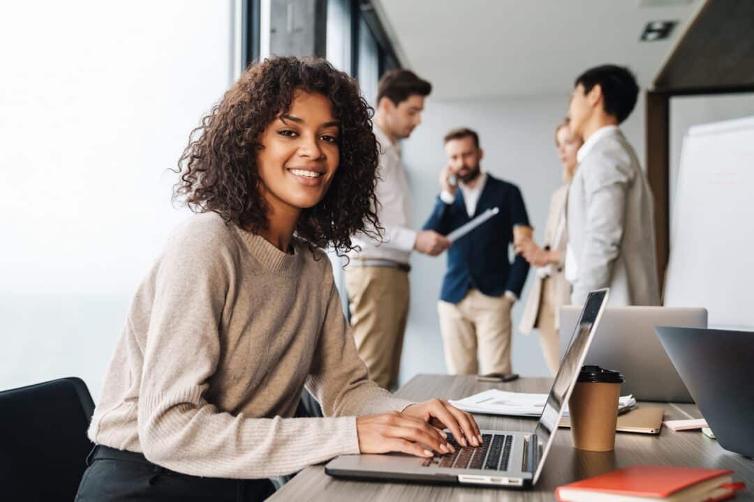 Imagem mostra uma jovem empresária sorrindo enquanto usa o computador. Atrás dela, estão alguns colegas de trabalho conversando.