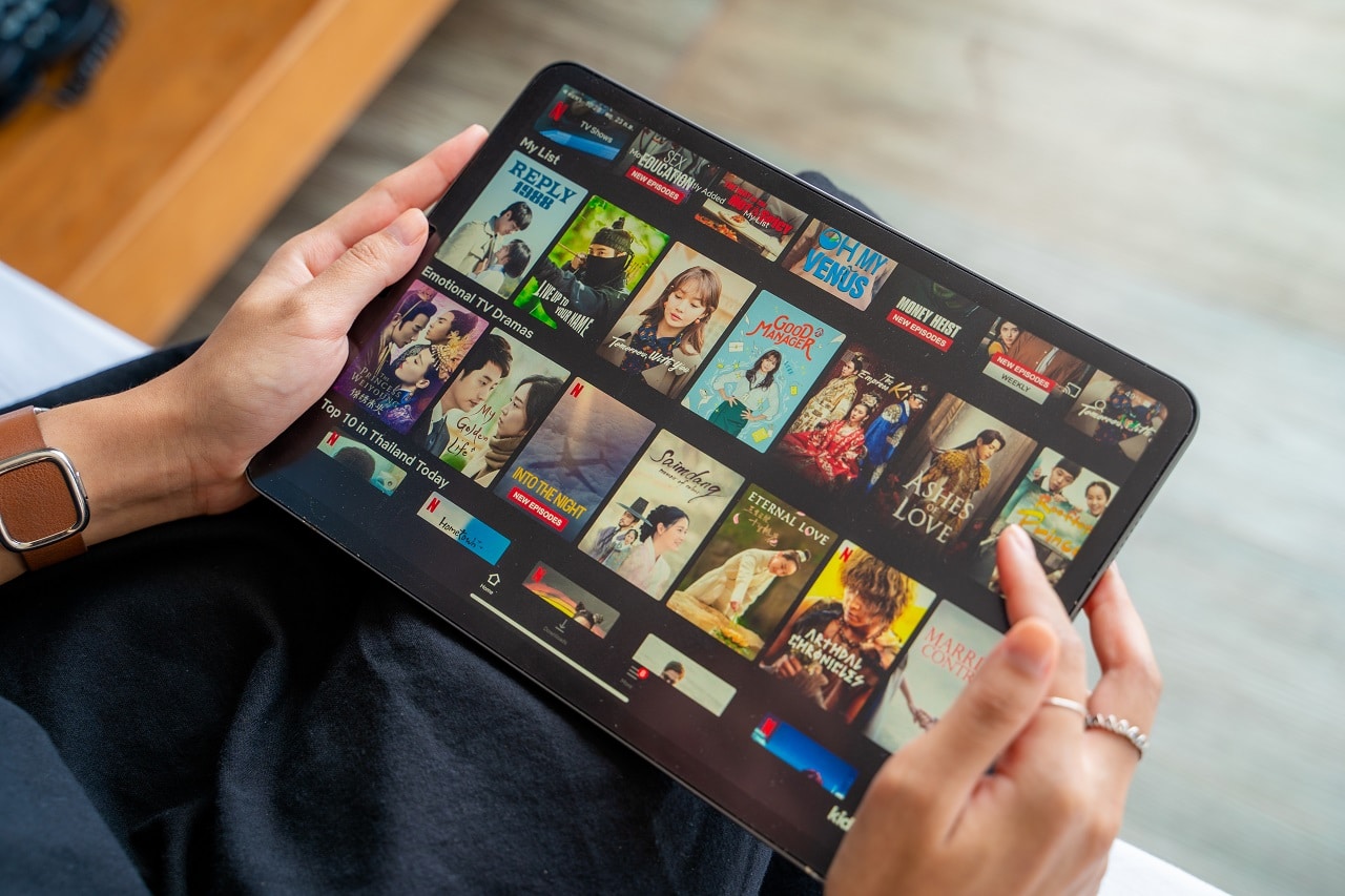 Imagem mostra duas mãos segurando um tablet que está aberto na página inicial da Netflix, com séries e filmes à disposição.