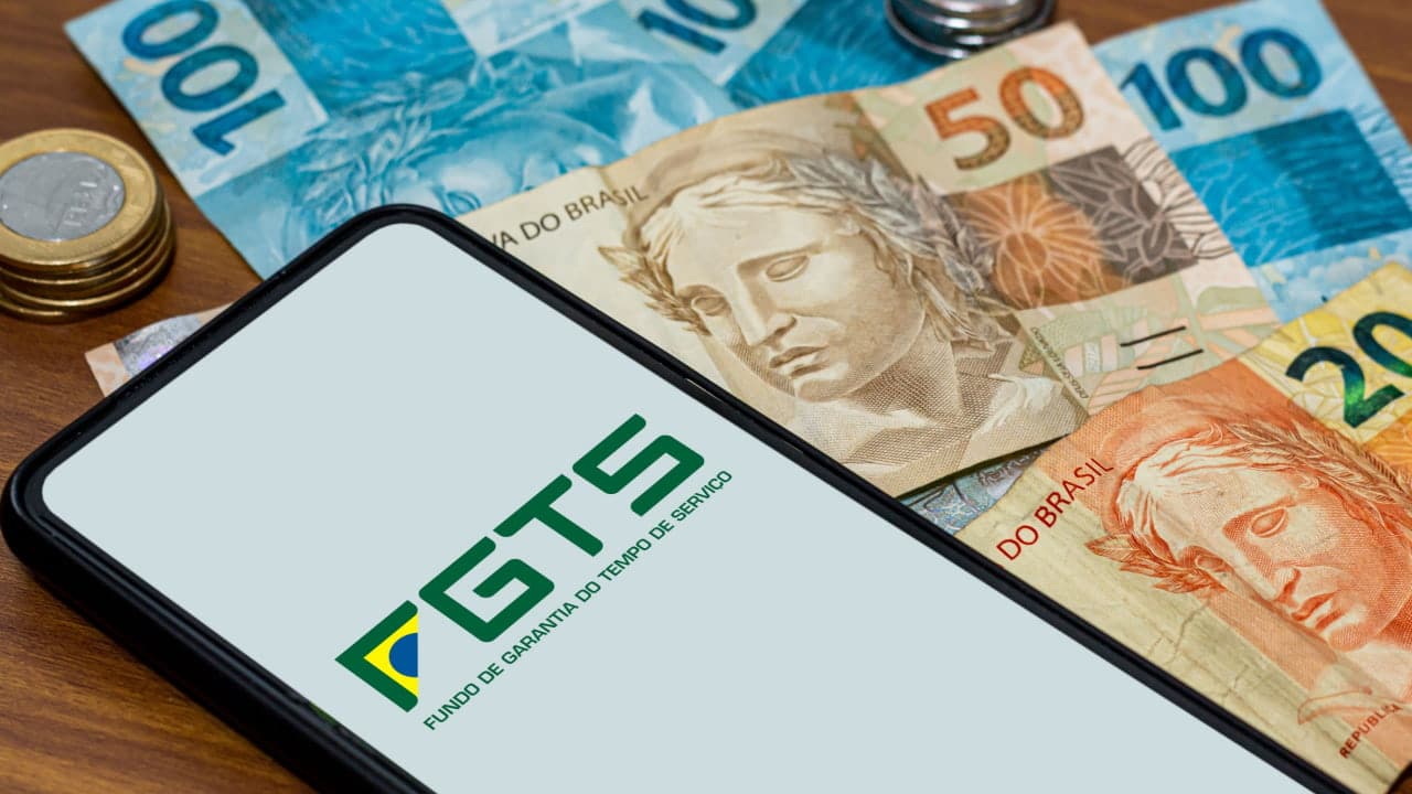Celular mostrando logo do FGTS (Fundo de Garantia do Tempo de Serviço) ao lado de notas de 50 reais, 100 reais e moedas
