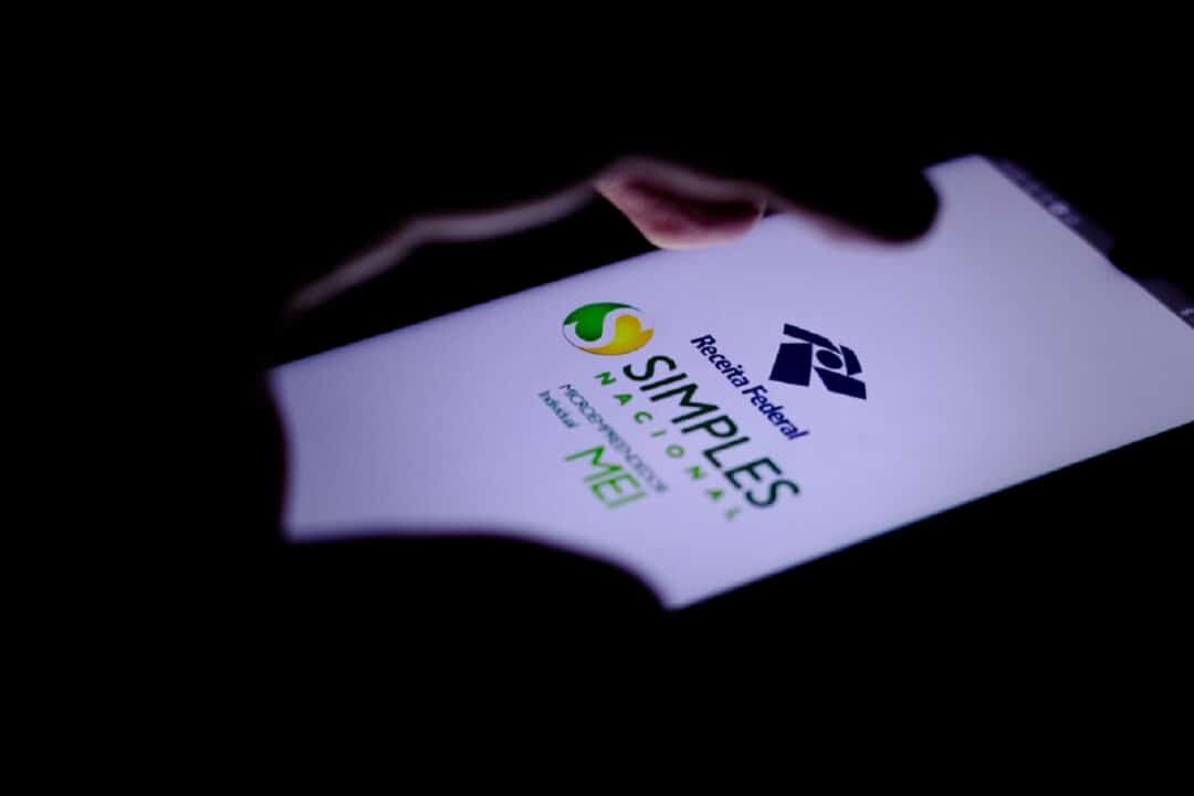 Mão tocando a tela de um celular que mostra a página inicial do aplicativo do Simples Nacional. O ambiente é escuro, com a única iluminação vindo da tela.