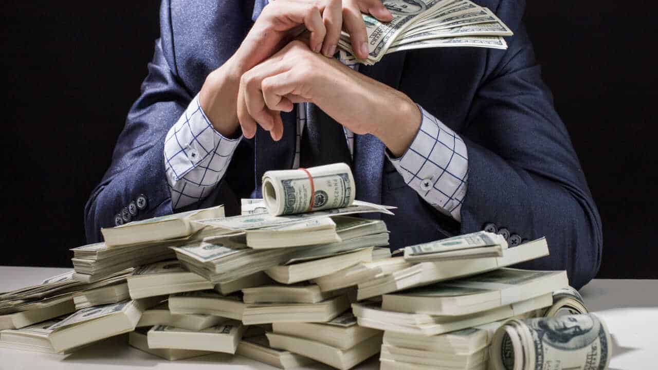 Homem segurando um monte de dinheiro e, sobre a mesa, diversas notas de dólares fortuna