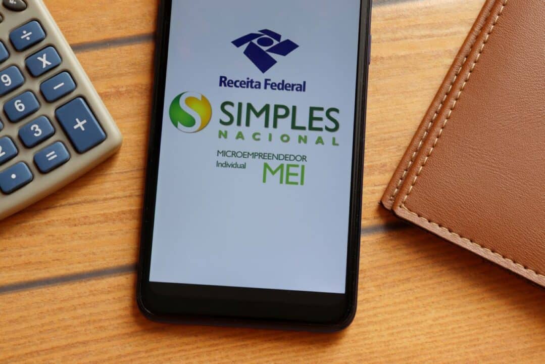 Dispostos em uma mesa de madeira, estão uma agenda, uma calculadora e um aparelho celular com o aplicativo Mais MEI aberto