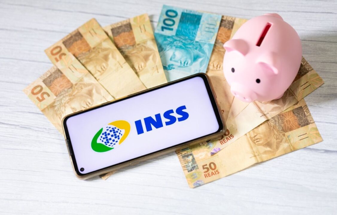 Na imagem se encontram um celular com o app o INSS aberto; ao lado, um cofre de porquinho e, embaixo, notas de 50 e 100 reais,
