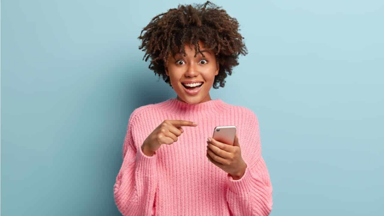 Mulher negra usando blusão rosa feliz apontando o dedo para o celular