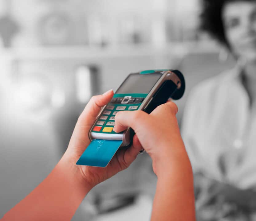Mãos digitando na maquininha de cartão. Ao fundo, em preto e branco, comerciante desfocada