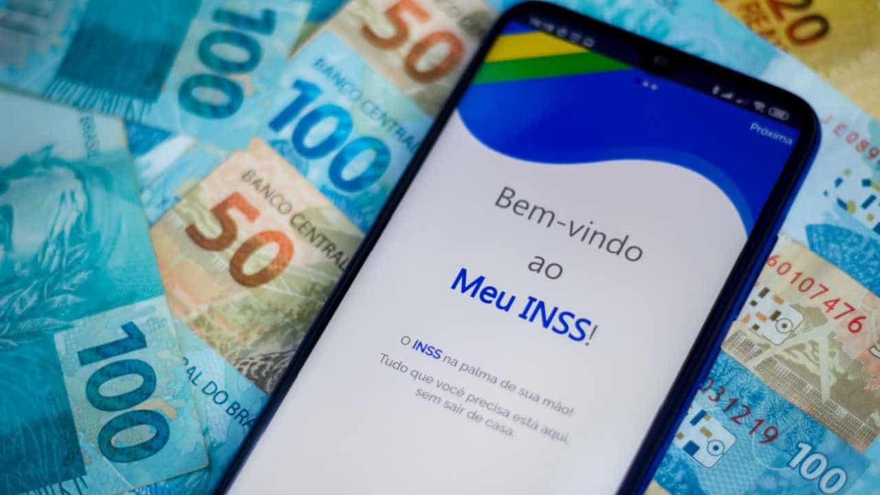 celular com app do INSS aberto em cima de notas de R$ 100 e R$ 50
