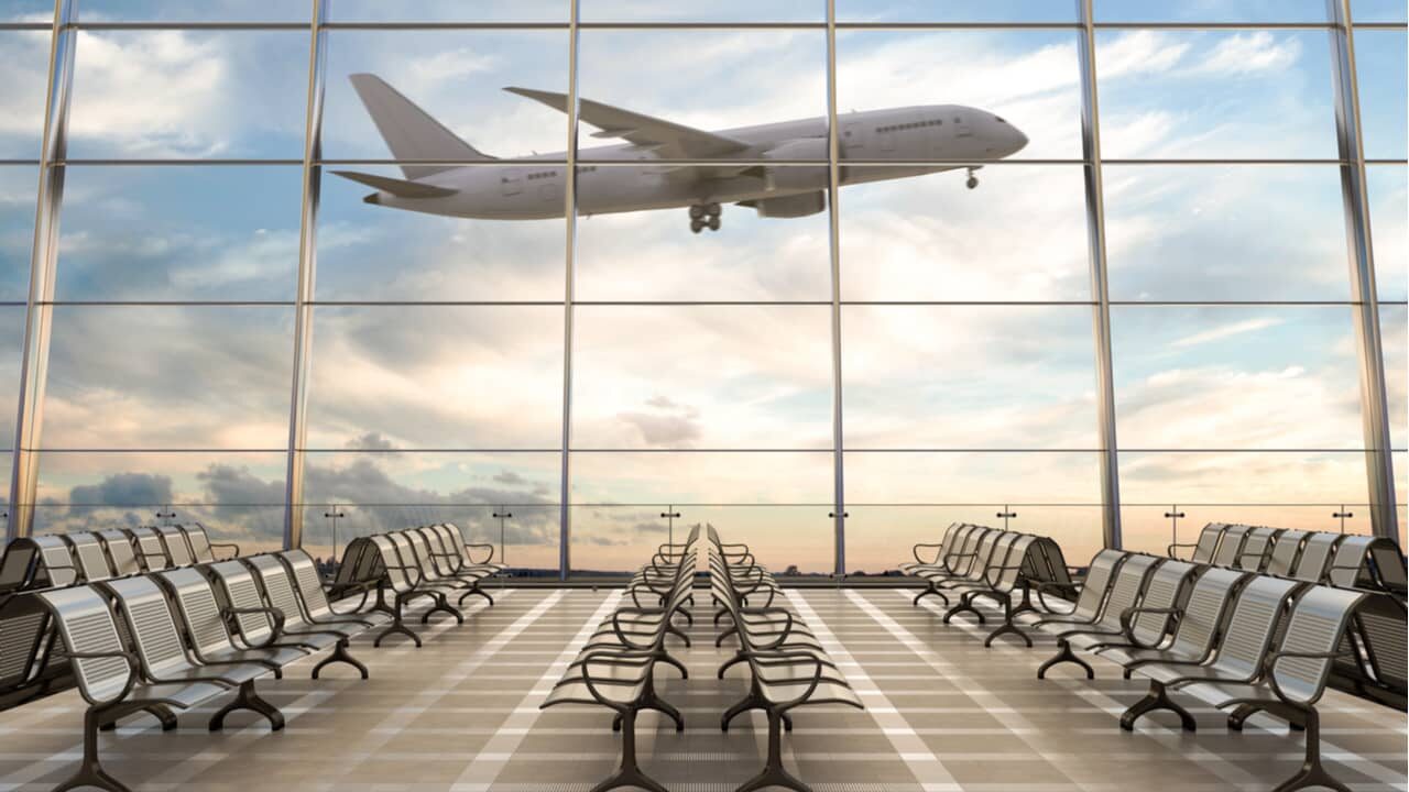 Foto tirada no interior de uma aeroporto focando em uma grande janela que dá vista para a decolagem de um grande avião.