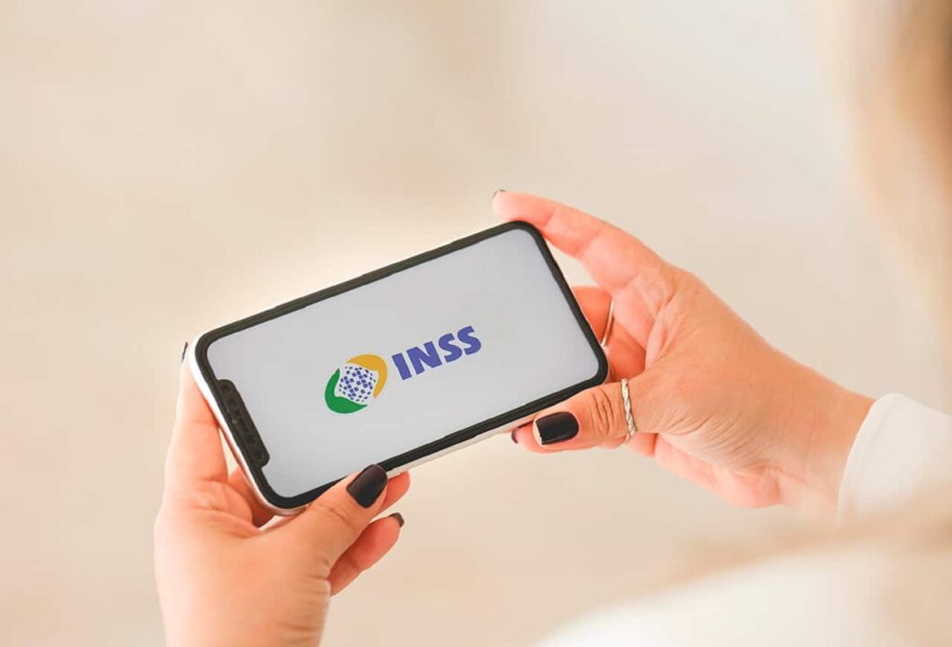 Imagem mostra duas mãos femininas segurando um celular. Na tela, é possível ver a logo do INSS.