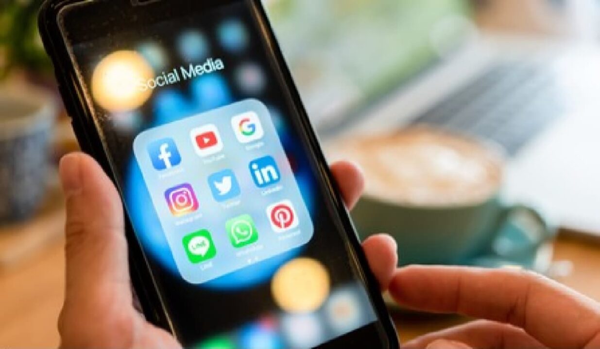 Foco em mão de pessoa segurando seu celular com uma pasta com redes sociais em evidência
