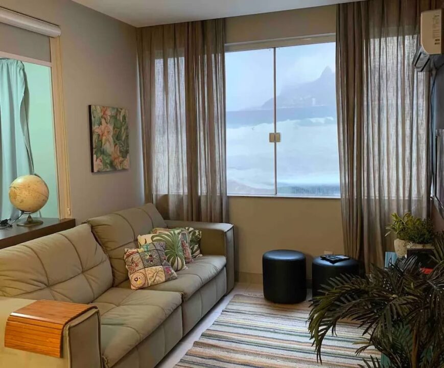 Sala de estar com sofá, tapete, plantas e janela com vista falsa para o mar.