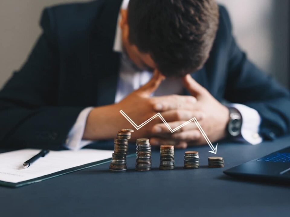 Imagem de empresário triste, na frente dele moedas empilhadas em forma de gráfico com uma seta indicando para baixo, representando o prejuízo do seu negócio.