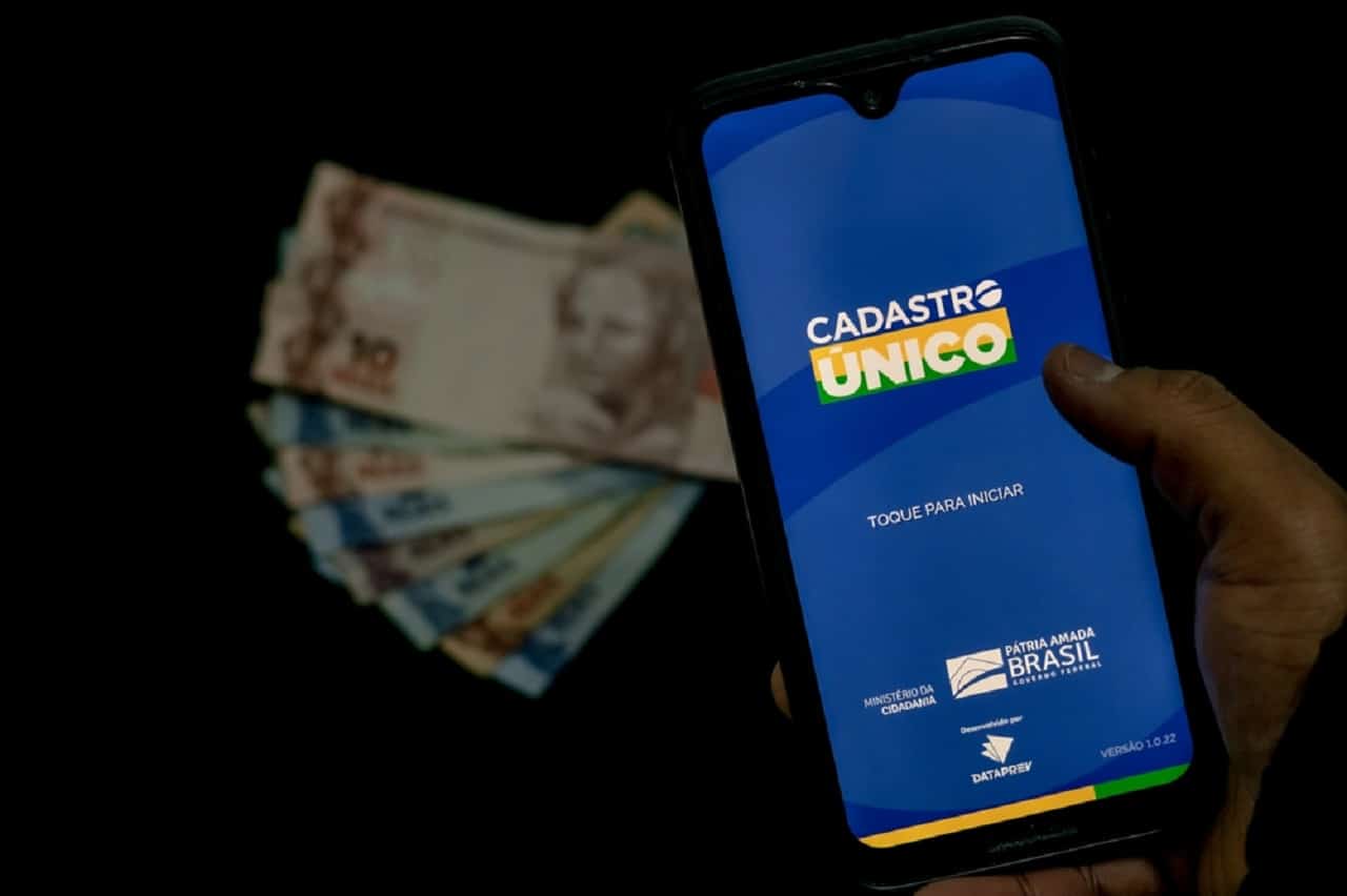 Imagem de um celular com o aplicativo do CadÚnico aberto. Ao fundo, algumas notas de R$10 e R$2 desfocadas.