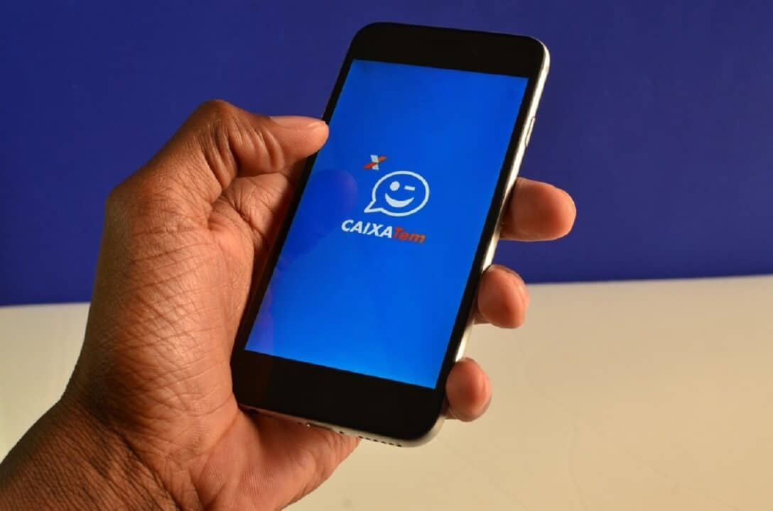 Imagem de uma mão segurando um celular com o aplicativo Caixa Tem aberto. O fundo está desfocado nas cores azul e bege.