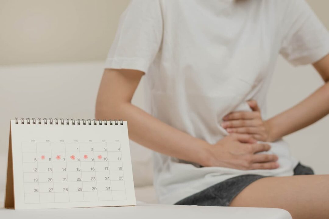 Calendário com dias marcados. Ao fundo uma pessoa com cólica. Imagem faz referência à licença menstrual