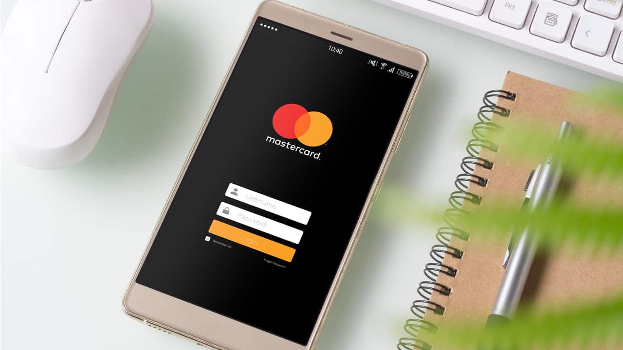 Celular com app do Mastercard aberto na tela de login. Ao redor, as espirais de um caderno, um mouse e um teclado de computador