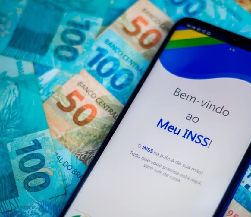 Celular exibindo o aplicativo Meu INSS colocado em cima de algumas notas de cem e cinquenta reais