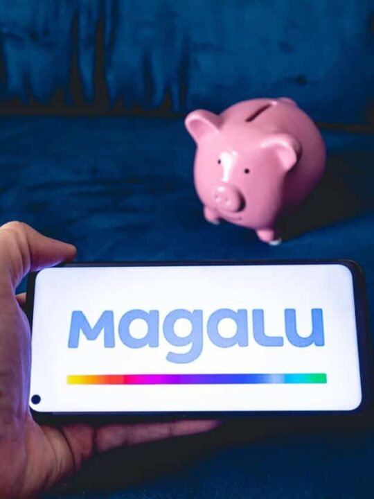 Mão segurando celular que mostra logo do Magalu (Magazine Luiza) em primeiro plano. Ao fundo, um cofrinho rosa em formato de porco.