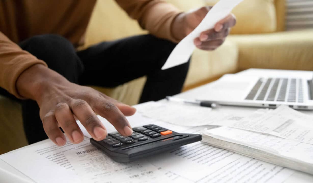 Homem fazendo contas na calculadora com recibos na mão e vários papéis espalhados na mesa a sua frente