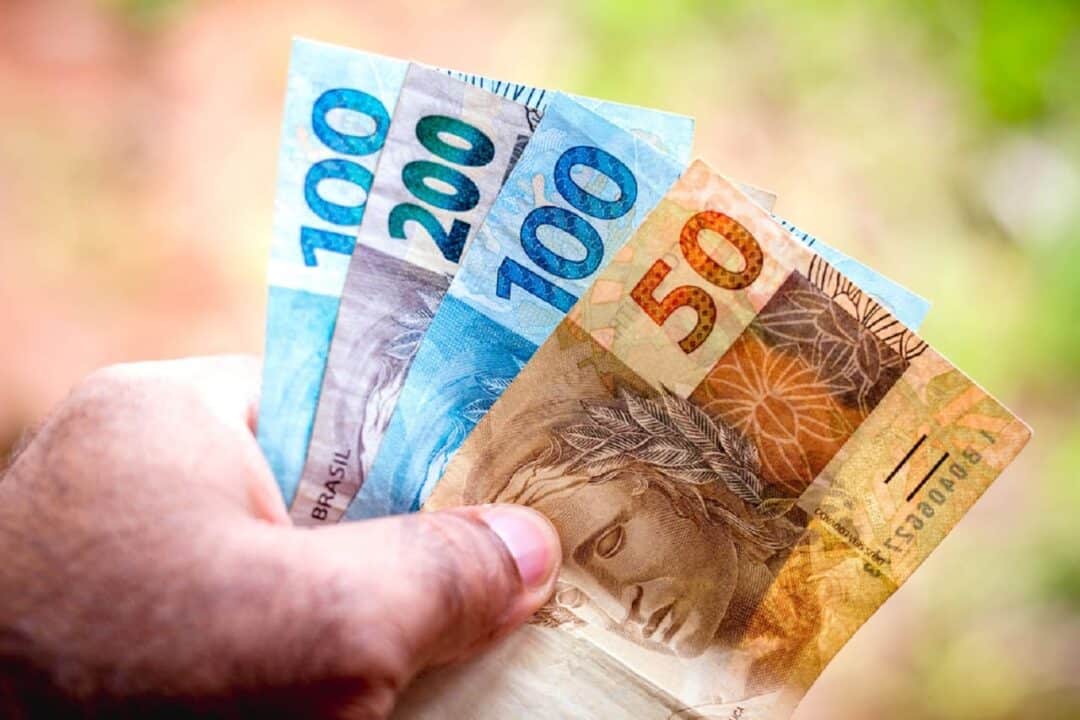 Imagem de uma mão segurando notas de R$200, R$100 e R$50 em formato de leque.