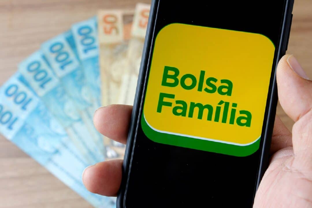 Pessoa segurando um celular com tela no logo do Bolsa Família. Abaixo se encontram notas de R$50 e R$100.