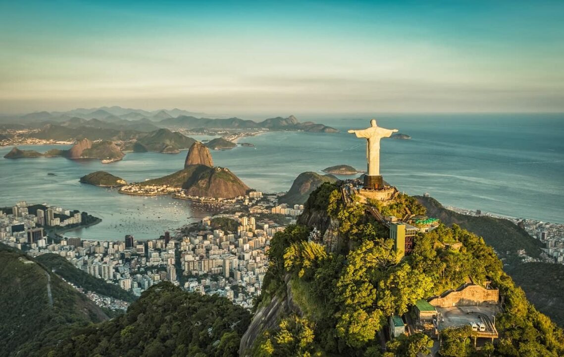 Imagem mostra uma visão aérea do Cristo Redentor, no Rio de Janeiro. Ao fundo, é possível ver um pedaço da cidade e da praia.