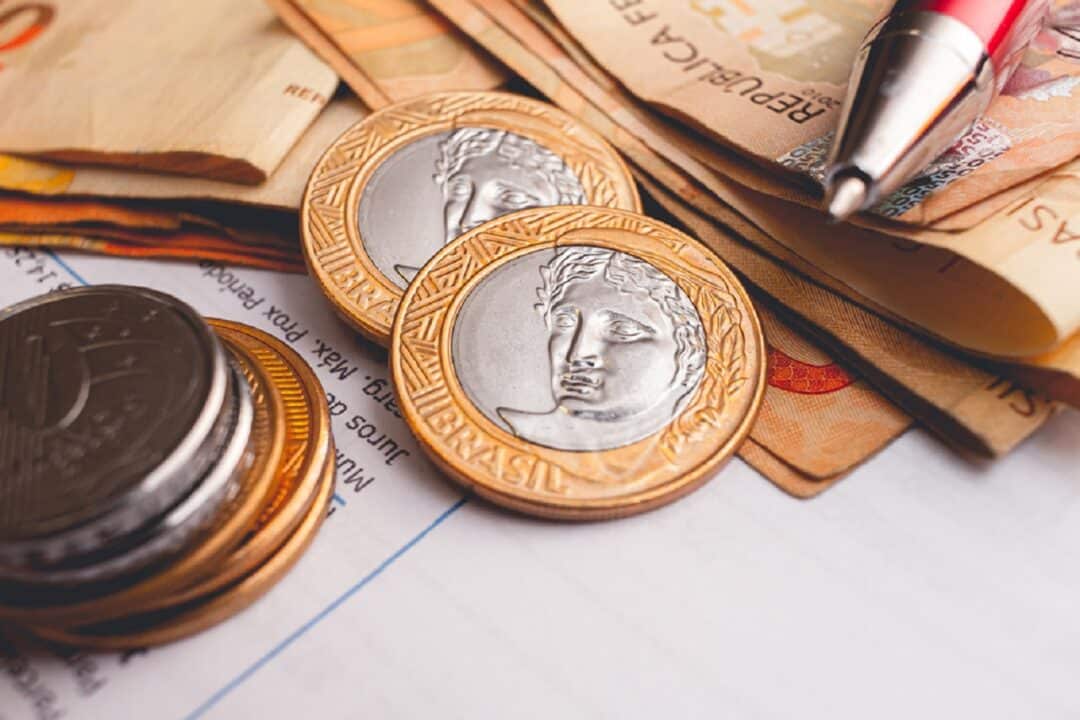 Sobre uma mesa estão notas de dinheiro, moedas, uma caneta e documentos.