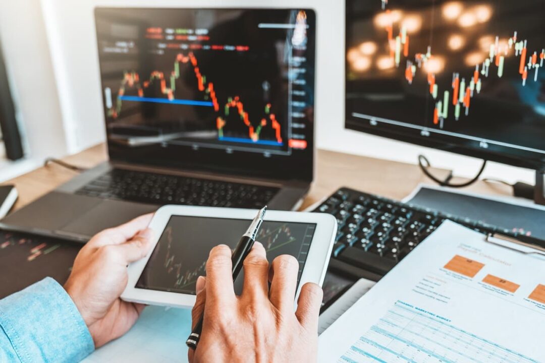 imagem de um homem analisando ações em gráficos financeiros exibidos em computador e tablet