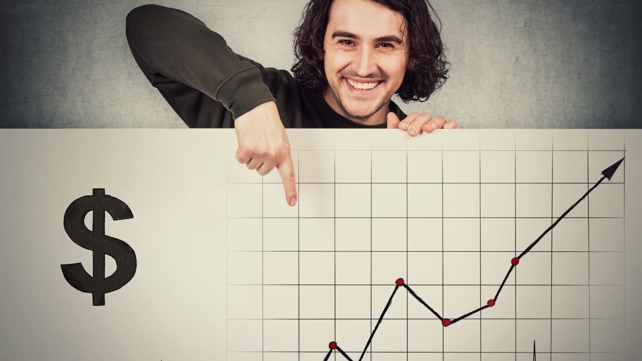 Homem feliz atrás de um quadro branco com esboços econômicos, apontando o dedo indicador para um gráfico financeiro crescente.
