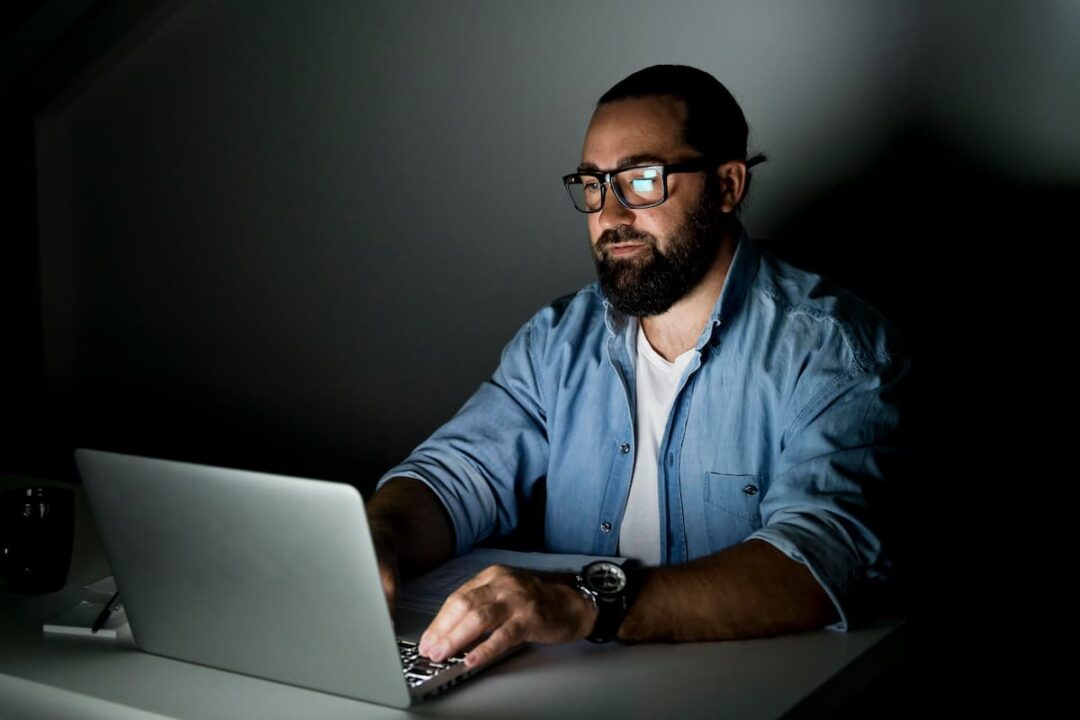 foto colorida de homem branco com barba e cabelo escuro, vestindo camiseta jeans e camisa branca, trabalhando no computador. Fundo cinza.