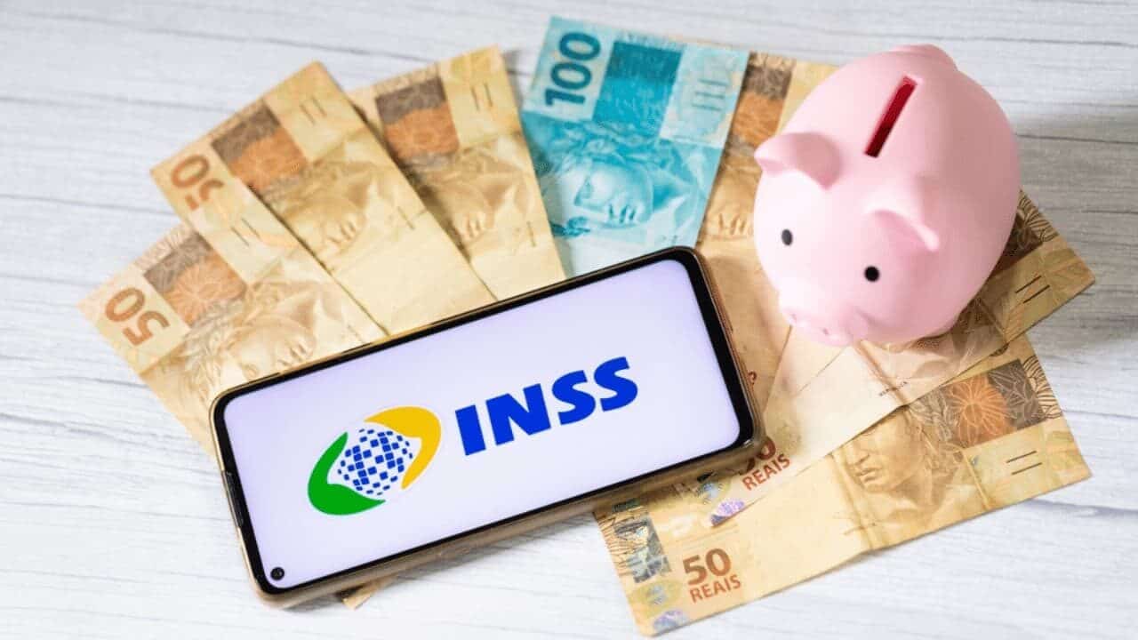 Um celular mostra na tela 'INSS', um cofre de porquinho rosa está ao lado e cédulas de 50 e 100 reais embaixo.