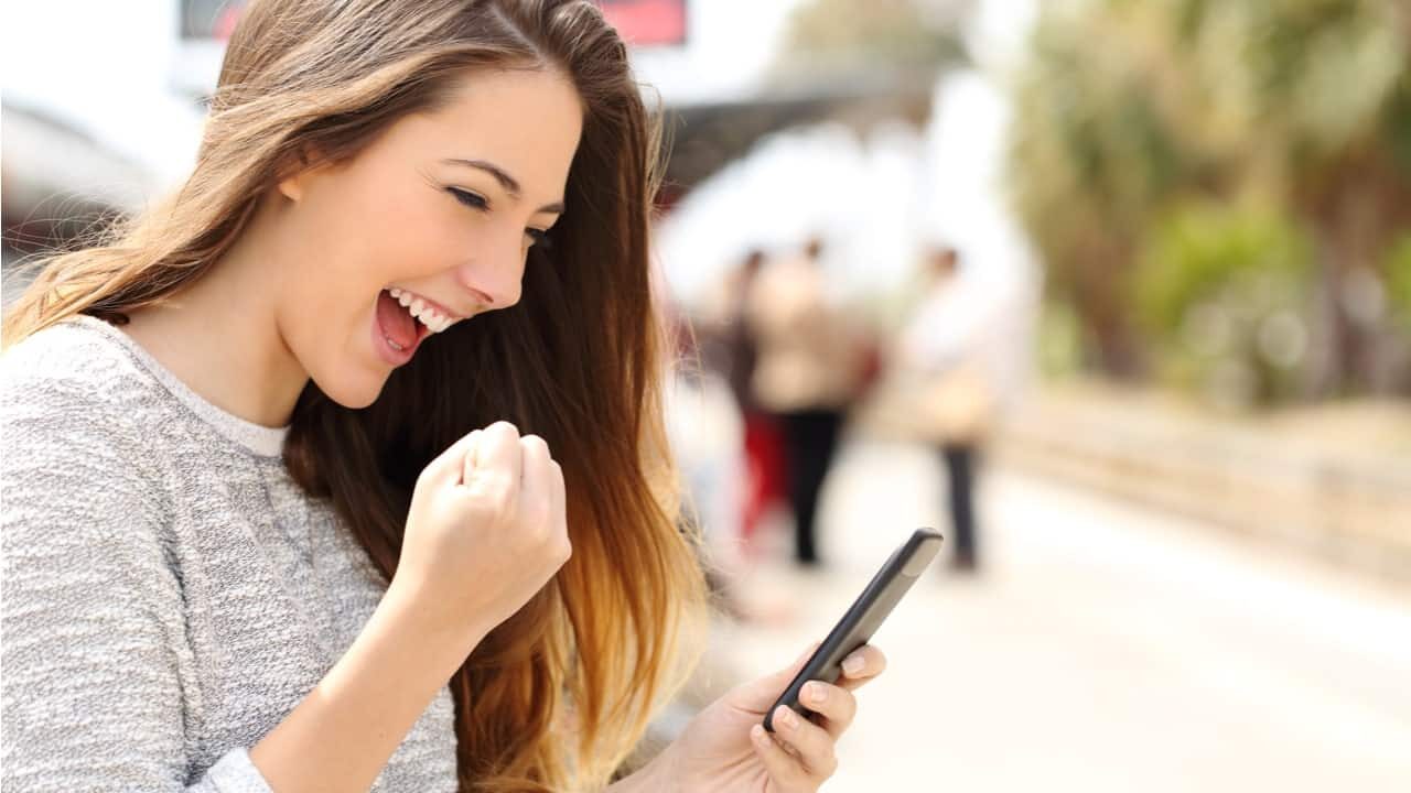 Uma jovem, branca, comemorando enquanto olha o celular.