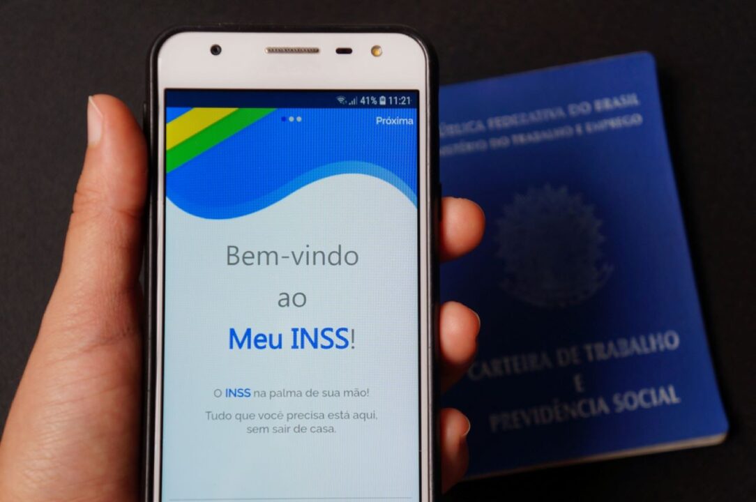 Mão femina segurando celular com o aplicativo Meu INSS aberto, com uma carteira de trabalho próxima