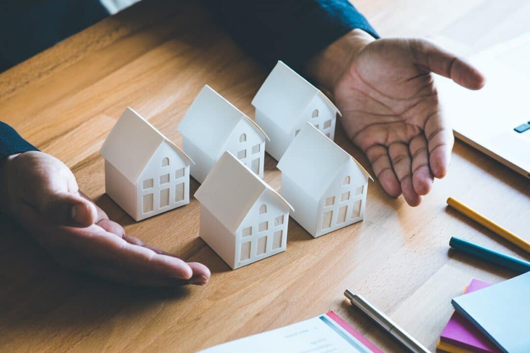 Imagem mostra uma mão masculina mostrando cinco maquetes brancas de casas sobre uma mesa